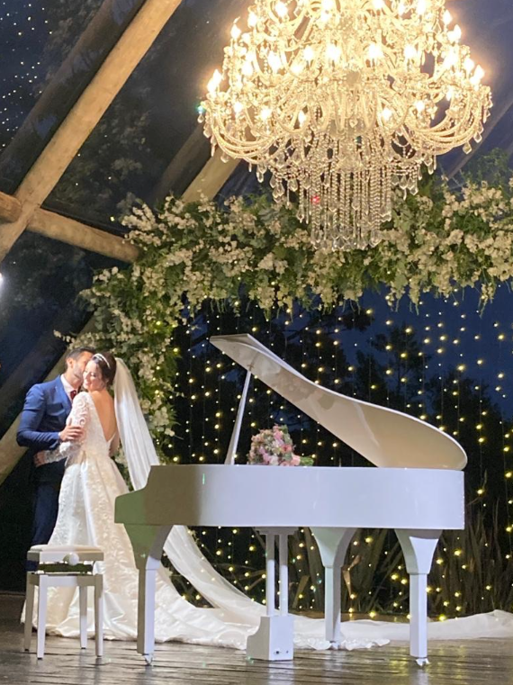 Casal junto ao piano branco, sob um lustre e flores em uma celebração de casamento