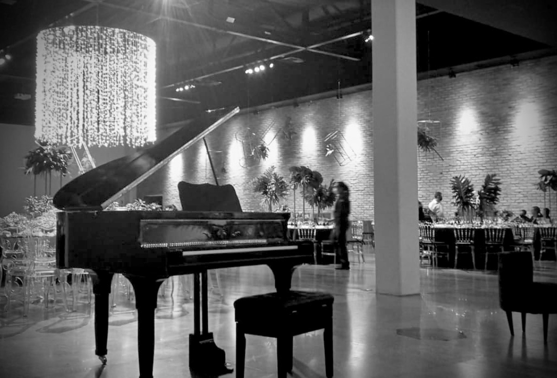 Piano preto em um espaço de eventos, fotografado em preto e branco, com palco e lustre ao fundo